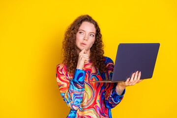Young woman with curly red hair holds a laptop against a bright yellow background, wearing stylish print clothes.