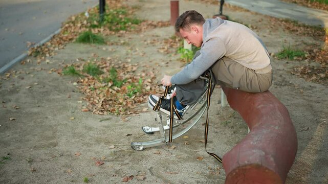 Man sitting on curved log in park, wearing light gray sweatshirt and olive-green pants, focused on adjusting straps of spring stilts with a determined expression, surrounded by fallen leaves
