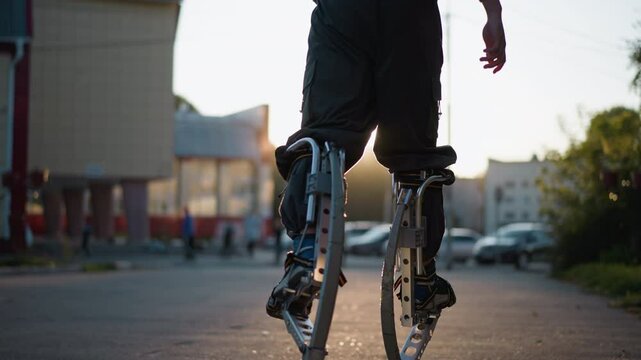 Back view of person walking on spring stilts during sunset wearing gray hoodie and pants. Background features blurred trees, suburban street, and warm sunlight creating soft shadows