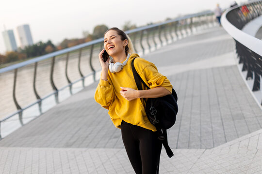 A joyful woman in a yellow sweater enjoys a phone call while walking along a riverside promenade on a sunny afternoon - Powered by Adobe