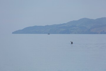 Obraz premium Kayaker paddles across calm water, serene landscape. Peaceful morning on the lake. NGARIMU BAY, THAMES, COROMANDEL PENINSULA, NEW ZEALAND
