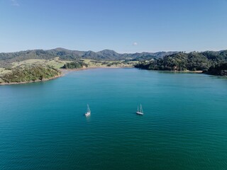Aerial view of two sailboats on a calm bay, surrounded by lush green hills and clear blue sky....