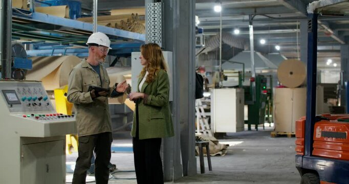 A man and a woman are talking standing in a paper mill. The man holds a tablet in his hand. They stand near the machine control panel