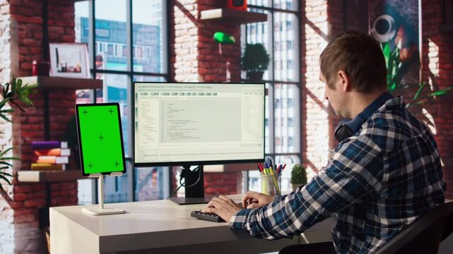 Programmer at desk typing code into computer, developing software programs, consulting green screen tablet. Man in home office fixing errors in application using chroma key device and PC, camera A