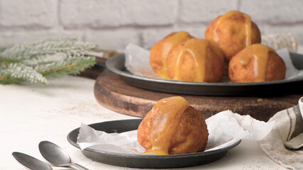 Golden-brown fried dough balls drizzled with a rich, creamy sauce are served on a rustic plate, evoking a cozy, festive atmosphere with a pine branch in the background