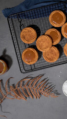 Caramel tarts on a cooling rack with a jar of caramel sauce, a bowl of salt, and a dried fern leaf on a dark surface, creating a rustic aesthetic