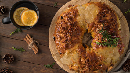 Festive wreath bread with nuts and honey glaze on a wooden board, accompanied by a cup of hot beverage with orange slices, cinnamon sticks, and pinecones