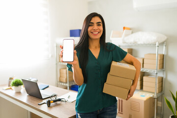 Cheerful latin woman holding packages and showing smartphone with blank screen in her small business store