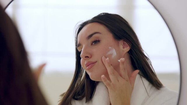 Cheerful young woman wearing bathrobe applying moisturizer cream on her face, enjoying her skincare routine in the bathroom