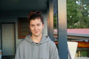 A casual portrait of a young woman wearing a grey hoodie, standing on a porch with a relaxed expression. The background features a mix of indoor and outdoor elements, including greenery and buildings