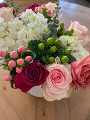 A beautiful floral arrangement featuring white hydrangeas, deep red and soft pink roses, green and pink berries, and eucalyptus stems, elegantly displayed on a wooden table in natural light.