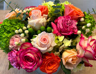 A vibrant bouquet showcasing pink, orange, and blush roses, white hydrangeas, green berries, white berries, and eucalyptus accents, arranged in a lush, colorful display on a wooden surface.