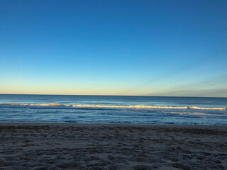 The sun sets over a calm ocean, casting soft golden light on gentle waves. The vast blue sky transitions into warm tones near the horizon, creating a serene and tranquil beach scene.