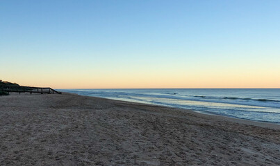 The beach extends toward a glowing horizon, where soft golden hues meet the tranquil blue sky at sunset. Gentle waves roll onto the sandy shore, creating a serene and expansive coastal scene.