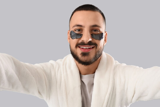 Young man in robe with eye patches taking selfie on grey background - Powered by Adobe