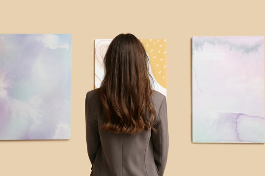 Young woman near paintings on beige wall in art gallery, back view