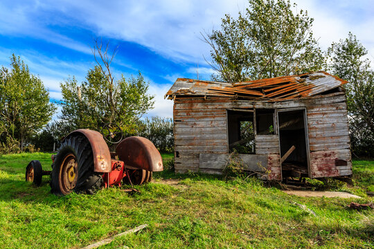 A dilapidated old farmhouse sits in a field with a red tractor in front of it