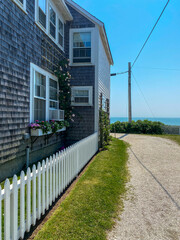 A coastal home with shingle siding, a white picket fence, blooming flower boxes, and a path leading to the ocean under a clear blue sky, embodying serene seaside charm.