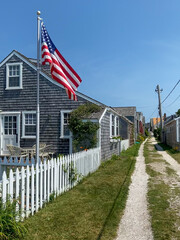 A quaint coastal cottage with shingle siding, an American flag, white picket fence, and a narrow gravel path, capturing a serene and patriotic seaside atmosphere under a clear blue sky.