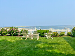 A peaceful backyard with white Adirondack chairs facing calm waters, vibrant hydrangeas, a white picket fence, and scattered boats under a bright blue sky, perfect for coastal relaxation.