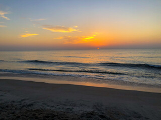 A peaceful sunrise on Jacksonville Beach, Florida, with gentle waves lapping the sandy shore. The sky is painted in soft orange and yellow hues as the sun rises above the horizon, creating a tranquil 