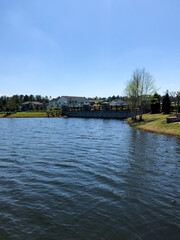 Fototapeta premium A calm lake stretches under a clear blue sky, bordered by grassy banks and a stone-lined bridge, with houses and trees in the distance, creating a peaceful suburban waterside scene.