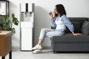 Beautiful woman drinking water on sofa near cooler in office