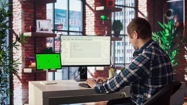 Man in home office looks at green screen tablet and computer, typing and executing programming code. Coding application used by IT developer writing and testing code using chroma key device, camera A