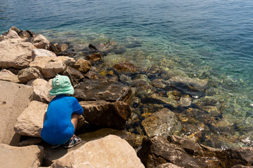 Young boy in green hat and blue swimsuit exploring rocky seashore by the clear blue water on a sunny summer day. Concept of childhood adventure, nature discovery, and outdoor activities by the sea.