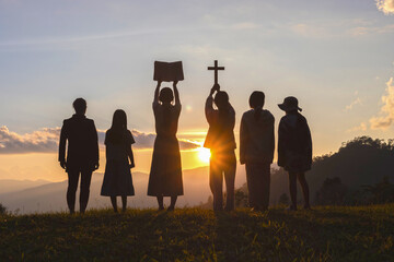 Silhouette of group children praying to the GOD while holding a crucifix symbol with bright sunbeam...