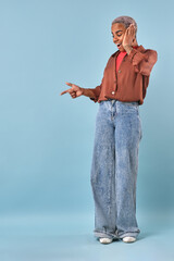 Young African American woman wearing a brown shirt and wide-legged jeans poses playfully against a soft blue background. Her joyful demeanor captivates the viewer as she gestures while smiling.