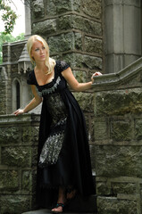 Sexy young blonde woman in vintage black dress or gown looking in the distance from the bottom stone staircase of castle like building