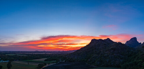 A panoramic view of dramatic mountain peaks silhouetted against a vibrant sunset sky, with fiery hues of red, orange, and pink blending into the fading blue. sunflowers fields around the mountains.