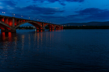 A bridge illuminated with red lights stretches over a river at dusk, with reflections on the water. The peaceful scene is captured during the blue hour, with no people in sight