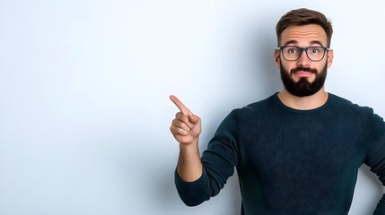 Fototapeta premium Confident young man with glasses and a beard gestures enthusiastically with his finger pointing to the side against a minimalist background, expressing excitement and curiosity.