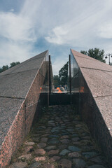 Obraz premium A solemn monument featuring an eternal flame between granite walls, with a cobblestone path leading through, symbolizing remembrance and respect against an open sky