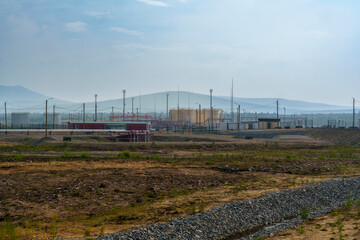 A remote construction site with a dirt road leading into the distance, surrounded by vegetation and mountains. Overcast sky and distant power lines create a rugged, industrial landscape with no people