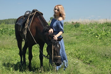 Abstract portrait of a romantic girl in a blue dress in a summer meadow, who feeds a black horse.