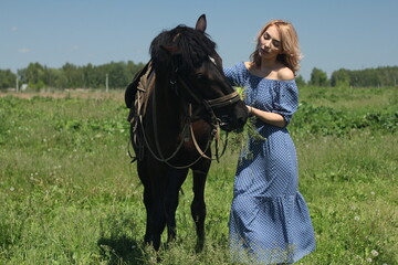 Obraz premium Abstract portrait of a romantic girl in a blue dress in a summer meadow, who feeds a black horse.