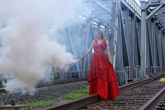 attractive girl in red dress on railroad near railway bridge gives the signal coming to the train by means of bright red objects and a smoke screen.	