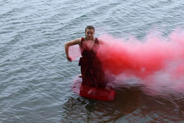 Fototapeta premium Abstract fashion portrait. Smoke under water. Young beautiful drowned woman with smoke grenade in red dress floating in the water after ship crush.