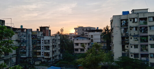 Urban apartment buildings stand closely together, silhouetted against a setting sun that casts a warm glow over the skyline. Lush greenery in the foreground contrasts with the concrete structures.