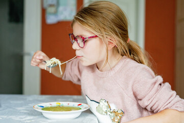 Funny little school girl eat chicken noodles soup with thin pasta. Happy preschool child eating fresh cooked healthy meal with noodles and vegetables at home, indoors.