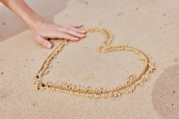 Person drawing a heart in the sand on the beach, with another hand reaching out to touch it