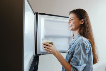 Smiling young woman with long hair holding a green smoothie in front of an open fridge, promoting healthy eating and lifestyle choices
