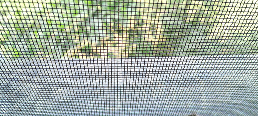 A fine mesh screen is in focus, with a blurry background of greenery and sunlight peeking through. The grid-like pattern creates a geometric effect, contrasting with the natural scene behind it.