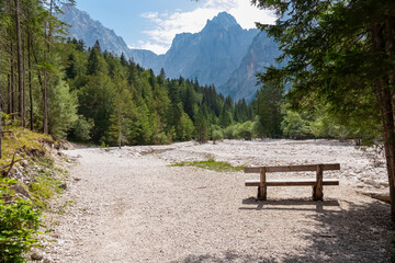 Fototapeta premium Wooden bench overlooking dried riverbank of Pisnica river winding through Krnica valley. Mountain peak Prisank, Julian Alps. Triglav national park, Vrsic pass, Slovenia. Wanderlust wild Slovenian Alps