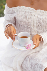 A close-up of a stylish woman wearing a white off-shoulder dress, holding a cup of tea on a saucer with a pink flower, enjoying a tranquil outdoor moment. Perfect for lifestyle, elegance, and