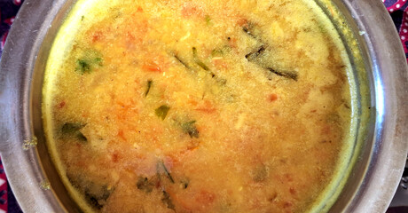 A steaming bowl of dal is filled with a mix of lentils, vegetables, and spices, showcasing a vibrant yellow color with hints of green and orange. 