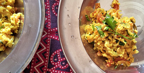 A plate of scrambled egg dish garnished with cilantro is placed on a vibrant, patterned cloth. The food appears colorful with visible pieces of tomatoes and herbs, served in a metallic dish.